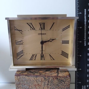 May include: A silver square clock with Roman numeral markings on a gold face. The clock reads "CITIZEN QUARTZ". The clock is sitting on a brown marble base.