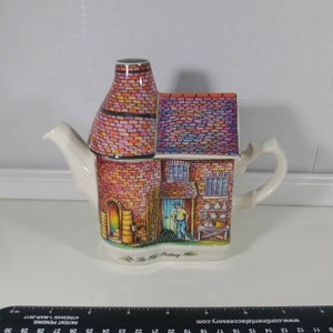 May include: A ceramic teapot shaped like a brick building with a chimney. The teapot is decorated with a colorful scene of a pottery shop with a man working inside. The text "The Old Pottery" is printed on the front of the teapot.