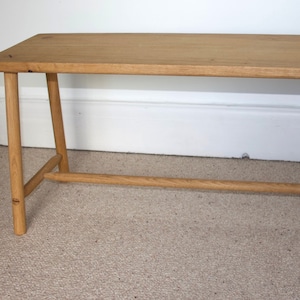 May include: A simple wooden bench with a natural finish. The bench has a rectangular top and four legs. The legs are angled and meet in the middle to form a support beam.