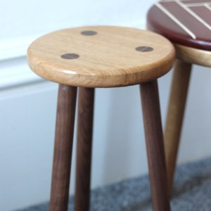 May include: A wooden stool with three dark brown legs and a light brown top. The top has three dark brown circles inlaid into the surface.