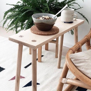 May include: A light wood bench with four legs and a natural wood finish. The bench is sitting on a white rug with a geometric pattern. A bowl of cereal and a mug are on top of the bench.