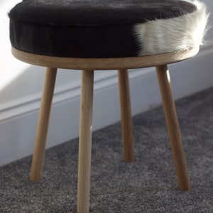 May include: A small, round stool with a black and white cowhide seat. The seat is supported by a light-colored wooden frame and three angled legs. The stool is a decorative furniture piece.