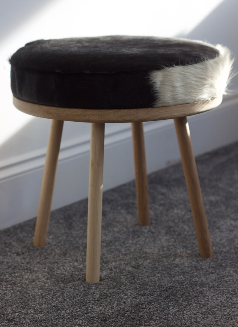 May include: A small, round stool with a black and white cowhide seat. The seat is supported by a light-colored wooden frame and three angled legs. The stool is a decorative furniture piece.