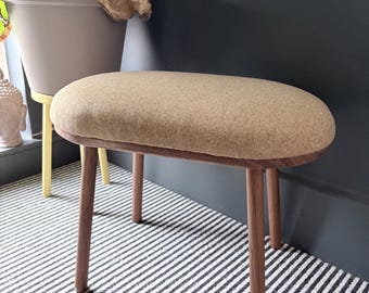May include: A small, oval-shaped stool with a light brown, textured seat and four wooden legs. The legs are a warm brown color. The stool is placed on a black and white striped rug.