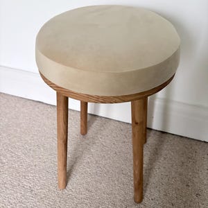 May include: A round stool featuring a smooth, light beige circular seat top and four tapered wooden legs. The legs are constructed from a medium-toned wood with a visible grain pattern, providing a sturdy base for the circular seat.