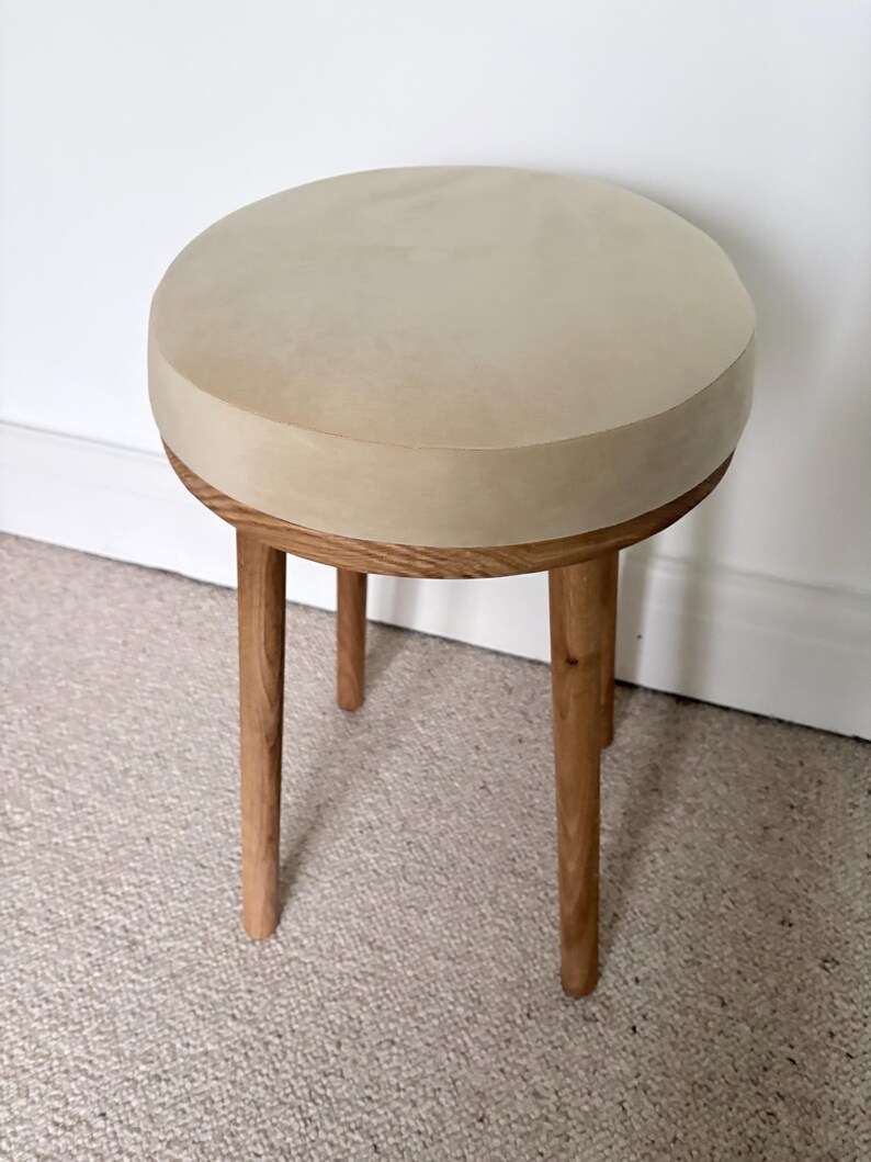 May include: A round stool featuring a smooth, light beige circular seat top and four tapered wooden legs. The legs are constructed from a medium-toned wood with a visible grain pattern, providing a sturdy base for the circular seat.