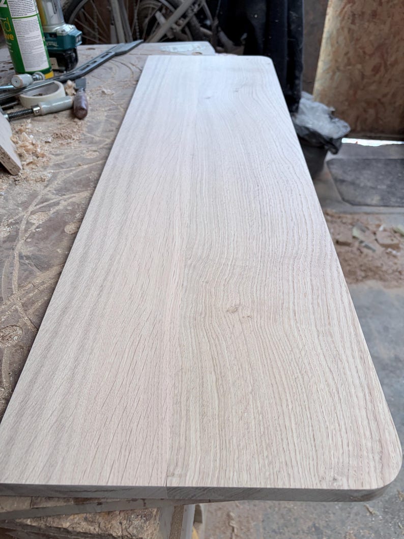 May include: A long, rectangular piece of light-coloured, unfinished wood board with a visible natural grain pattern. The board features smooth, flat surfaces and rounded corners on one end, showcasing the raw texture of the timber.