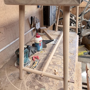 May include: A light-colored wooden stool featuring a rectangular top supported by four cylindrical legs. The legs are connected by a horizontal cylindrical stretcher bar, and the joints are secured with small, dark wooden wedges visible at the top.