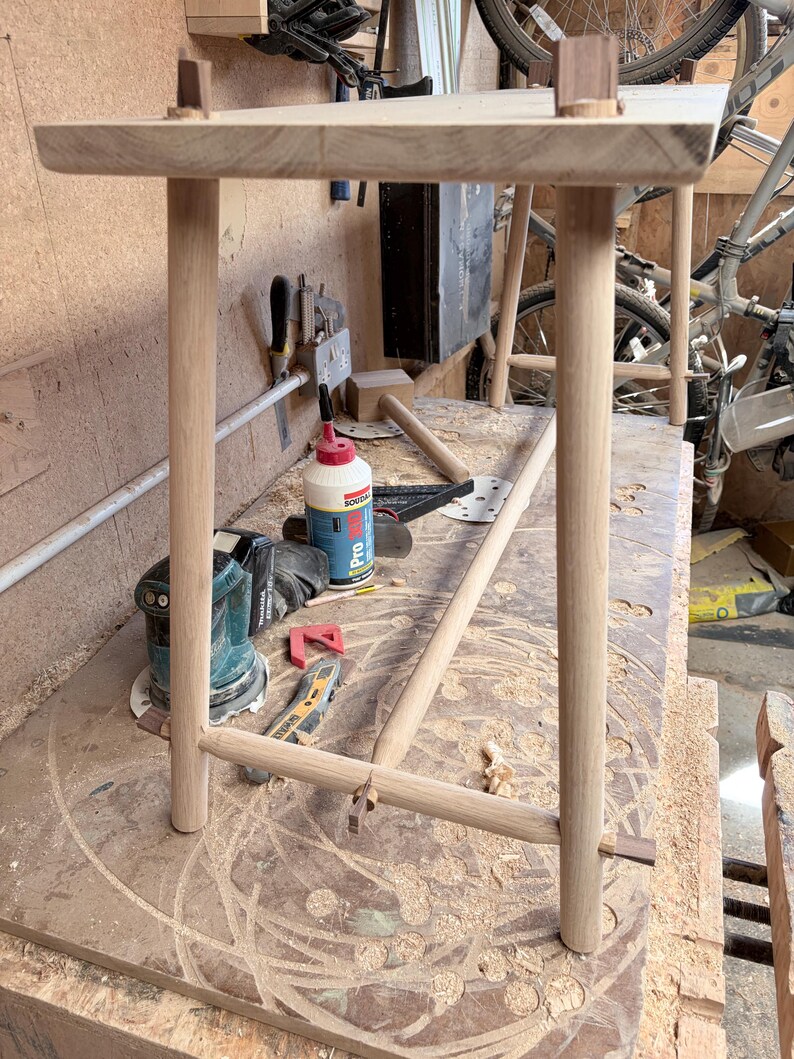 May include: A light-colored wooden stool featuring a rectangular top supported by four cylindrical legs. The legs are connected by a horizontal cylindrical stretcher bar, and the joints are secured with small, dark wooden wedges visible at the top.