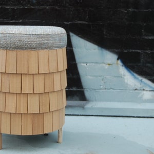 May include: A wooden stool with a round, gray fabric seat. The stool has a unique design with multiple layers of wood that resemble shingles.
