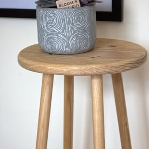 May include: A wooden stool with three legs and a round top. A gray planter with a succulent plant is sitting on the stool. The planter has a label that says "Bloomin'  Fabulous".