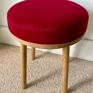 May include: A round stool featuring a deep red velvet upholstered seat cushion. The base is constructed from light-colored natural wood, consisting of a circular frame and three sturdy, cylindrical wooden legs that provide stable support.
