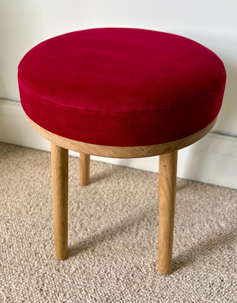May include: A round stool featuring a deep red velvet upholstered seat cushion. The base is constructed from light-colored natural wood, consisting of a circular frame and three sturdy, cylindrical wooden legs that provide stable support.