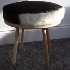 May include: A round stool with a black and white cowhide seat and light wood legs. The seat is a mix of black and white fur. The stool has three angled legs made of light-colored wood, creating a simple, natural aesthetic.