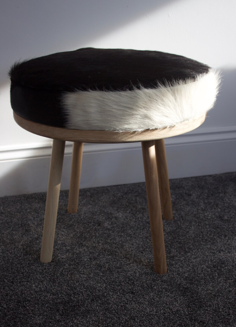 May include: A round stool with a black and white cowhide seat and light wood legs. The seat is a mix of black and white fur. The stool has three angled legs made of light-colored wood, creating a simple, natural aesthetic.