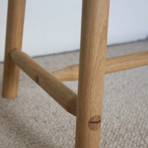 May include: A light brown wooden bench featuring cylindrical legs and horizontal support stretchers. The joinery displays visible circular wooden dowels with a dark accent line, connecting the legs to the horizontal support bars.