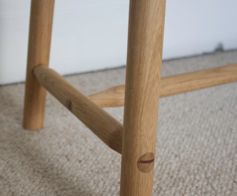 May include: A light brown wooden bench featuring cylindrical legs and horizontal support stretchers. The joinery displays visible circular wooden dowels with a dark accent line, connecting the legs to the horizontal support bars.