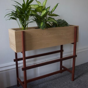 Könnte beinhalten: Ein hölzerner Pflanzenkasten mit drei Pflanzen, der auf einem roten Metallständer mit zwei Regalen steht. Der Pflanzenkasten ist hellbraun und der Ständer ist dunkelrot.