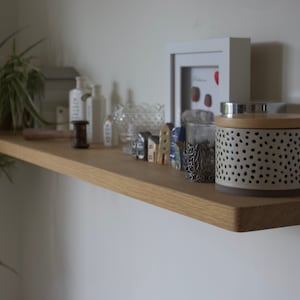 May include: A light-toned wooden floating shelf holding a potted spider plant, white bottles, a glass bowl, small house-shaped figurines, a silver filigree cup, and a cylindrical container with a black-spotted pattern and wooden lid.