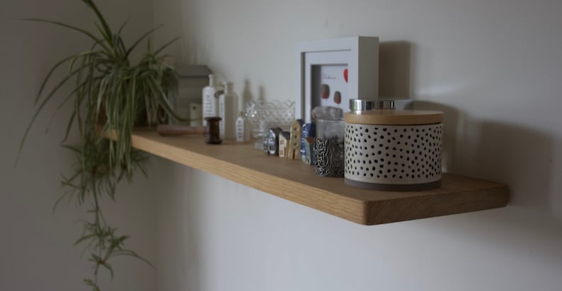 May include: A light-toned wooden floating shelf holding a potted spider plant, white bottles, a glass bowl, small house-shaped figurines, a silver filigree cup, and a cylindrical container with a black-spotted pattern and wooden lid.