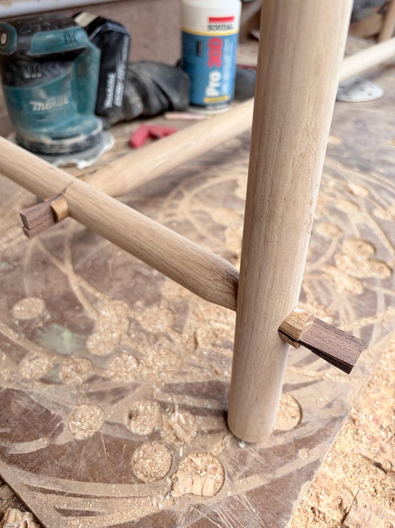 May include: A light-colored wooden furniture frame featuring cylindrical legs and horizontal crossbars connected by dark brown wooden wedge joinery. The structure displays smooth, sanded surfaces with visible wood grain and precise mortise and tenon joints.