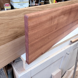 May include: Two rectangular wooden boards positioned upright. The board in the foreground is a deep reddish-brown wood with a smooth finish and rounded edges. The board behind it is a lighter, golden-tan wood with a visible grain pattern.