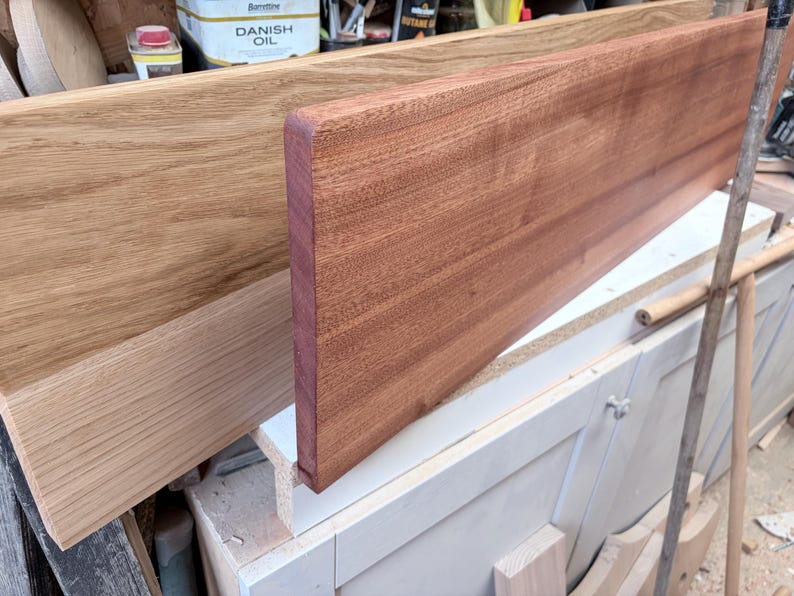 May include: Two rectangular wooden boards positioned upright. The board in the foreground is a deep reddish-brown wood with a smooth finish and rounded edges. The board behind it is a lighter, golden-tan wood with a visible grain pattern.