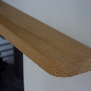 May include: A light brown oak wood mantel shelf with a smooth, flat top surface and a rounded front edge. The shelf is mounted horizontally against a plain white wall, showcasing the natural wood grain texture and light tan colour.