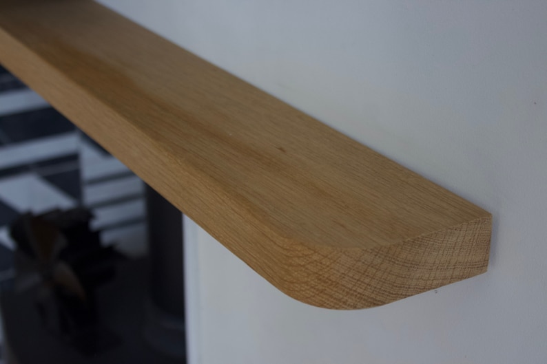 May include: A light brown oak wood mantel shelf with a smooth, flat top surface and a rounded front edge. The shelf is mounted horizontally against a plain white wall, showcasing the natural wood grain texture and light tan colour.