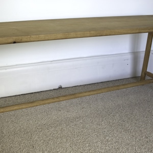 May include: A rectangular light brown wooden bench featuring a flat top surface and four cylindrical legs connected by a horizontal support bar running between the front and back legs on each side, showcasing a natural wood grain finish.