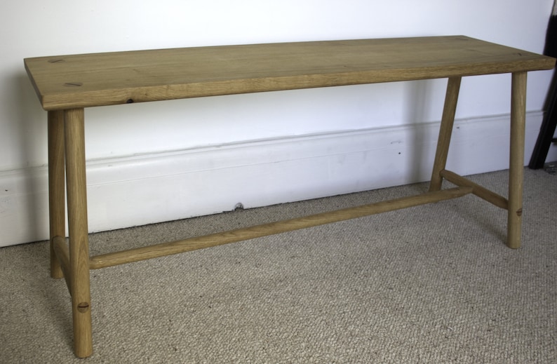 May include: A rectangular light brown wooden bench featuring a flat top surface and four cylindrical legs connected by a horizontal support bar running between the front and back legs on each side, showcasing a natural wood grain finish.