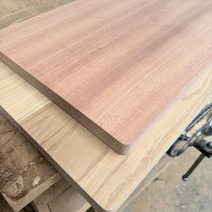 May include: Two rectangular wooden boards stacked on top of each other. The top board is a reddish-brown wood with a smooth surface, while the bottom board is a lighter, pale tan wood with a visible grain pattern and rounded corners.