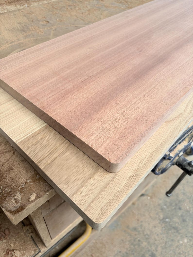 May include: Two rectangular wooden boards stacked on top of each other. The top board is a reddish-brown wood with a smooth surface, while the bottom board is a lighter, pale tan wood with a visible grain pattern and rounded corners.