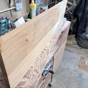 May include: A rectangular light brown wooden board with a smooth, rounded left edge and visible natural wood grain patterns. The board is positioned at an angle, showcasing its flat surface and thickness in a workshop environment.