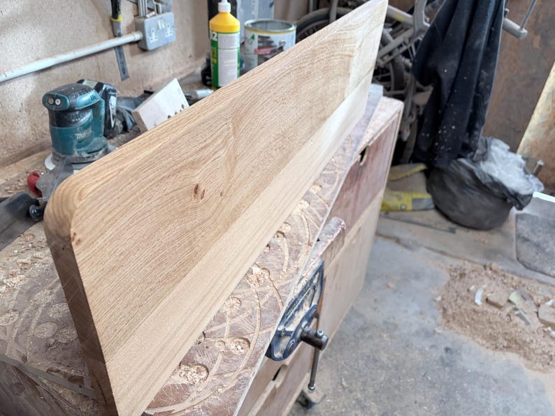 May include: A rectangular light brown wooden board with a smooth, rounded left edge and visible natural wood grain patterns. The board is positioned at an angle, showcasing its flat surface and thickness in a workshop environment.