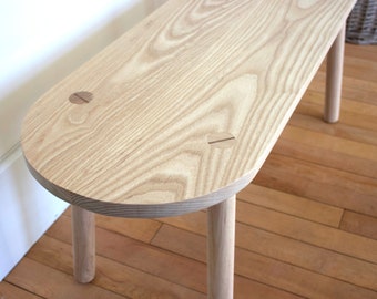May include: A light wood bench with a rounded top and three legs. The bench has a natural wood finish and two wooden dowels on the top.