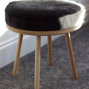 May include: A small, round stool with a natural wood frame and legs. The seat is upholstered with a black and white cowhide pattern. The stool has three angled legs and is suitable for a living room or bedroom.