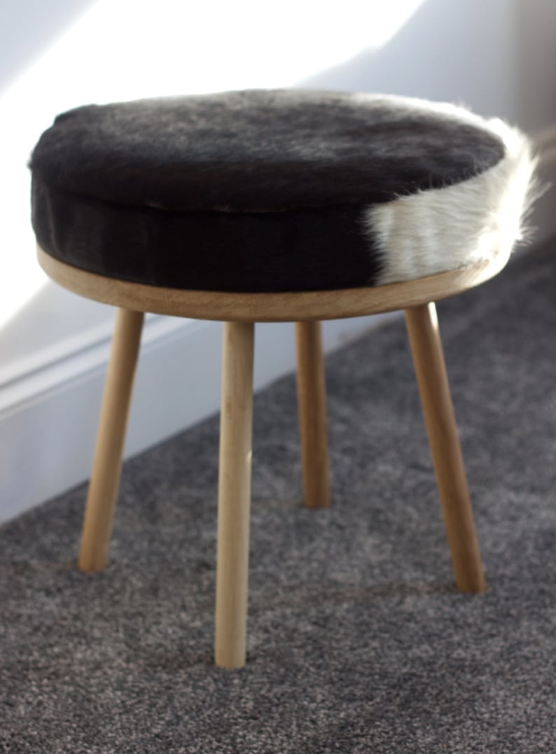May include: A small, round stool with a natural wood frame and legs. The seat is upholstered with a black and white cowhide pattern. The stool has three angled legs and is suitable for a living room or bedroom.