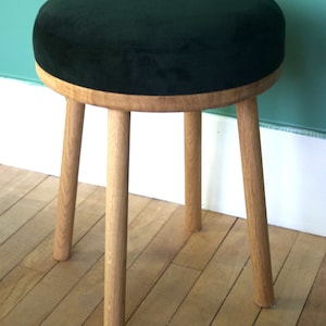 May include: A round stool featuring a dark green velvet upholstered seat cushion set upon a light-colored natural wood frame. The stool is supported by four cylindrical wooden legs that extend downwards from the circular wooden base.