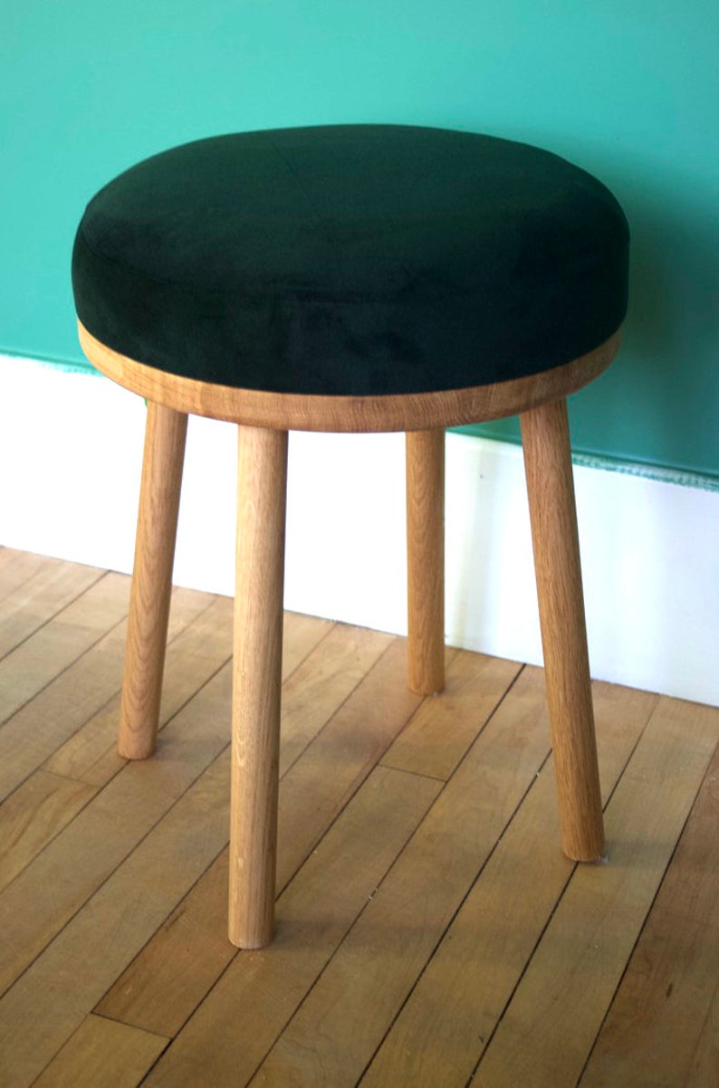 May include: A round stool featuring a dark green velvet upholstered seat cushion set upon a light-colored natural wood frame. The stool is supported by four cylindrical wooden legs that extend downwards from the circular wooden base.