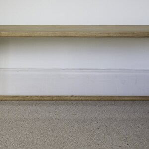 May include: A light brown wooden bench featuring a rectangular seat with rounded edges, supported by four splayed legs connected by a horizontal stretcher bar near the base. The wood grain is visible throughout the entire structure of the bench.