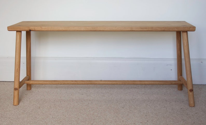 May include: A light brown wooden bench featuring a rectangular seat with rounded edges, supported by four splayed legs connected by a horizontal stretcher bar near the base. The wood grain is visible throughout the entire structure of the bench.