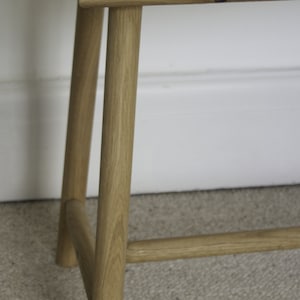 May include: A light brown wooden bench featuring a rectangular top with visible circular joinery details. The bench has four cylindrical legs connected by horizontal support bars, showcasing a natural wood grain finish throughout the structure.