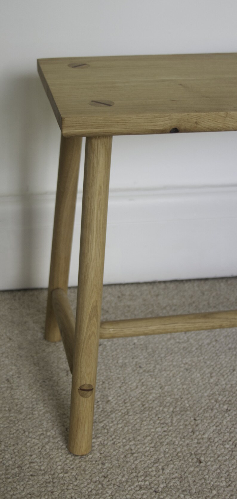 May include: A light brown wooden bench featuring a rectangular top with visible circular joinery details. The bench has four cylindrical legs connected by horizontal support bars, showcasing a natural wood grain finish throughout the structure.