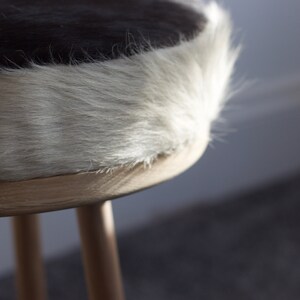 May include: A close-up of a small, round stool with a cowhide seat. The seat features a black and white pattern, with a fluffy, light-colored edge. The stool has three wooden legs.