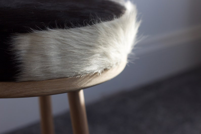 May include: A close-up of a small, round stool with a cowhide seat. The seat features a black and white pattern, with a fluffy, light-colored edge. The stool has three wooden legs.