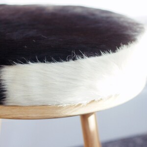 May include: A round stool with a cowhide seat. The seat is black and white, with a natural wood base and three legs. The stool is a decorative piece of furniture.