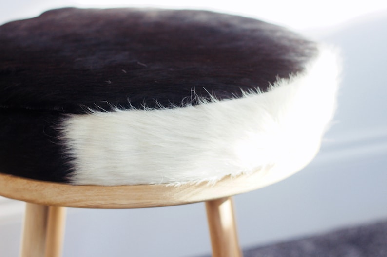 May include: A round stool with a cowhide seat. The seat is black and white, with a natural wood base and three legs. The stool is a decorative piece of furniture.