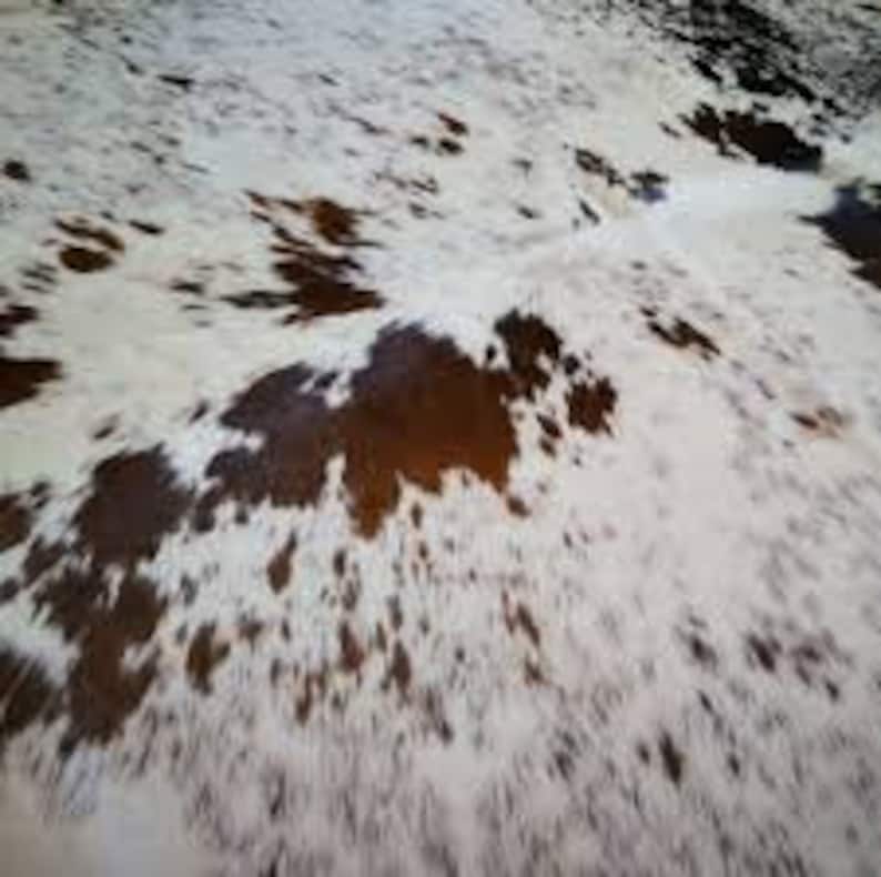 May include: A close-up of a cowhide rug with a natural pattern. The rug features a mix of white, brown, and black spots, creating a unique and organic design. The texture appears soft and inviting.