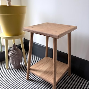 May include: A light brown wooden side table with a square top and a lower square shelf, supported by four straight legs. The table features a visible wood grain pattern and joinery details on the top surface, constructed from natural timber.
